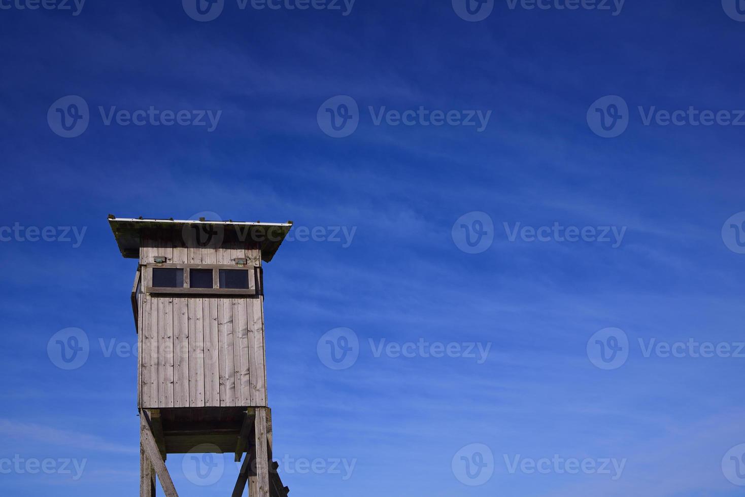 A weathered wooden stand for hunters stands on wooden posts and has a narrow window. The high stand is at the bottom of the picture against a blue sky with clouds in Bavaria photo