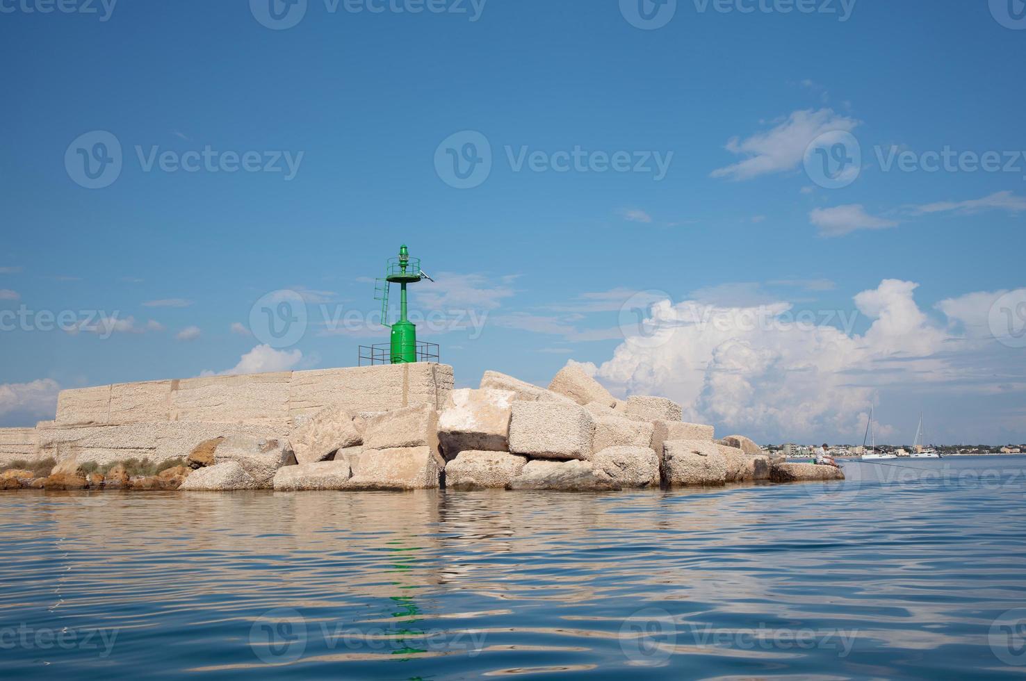 A green metal signal tower stands on the shore of the Mediterranean Sea. The tower stands on many blocks of stone. In the background the blue sky reflected in the water and the waves. photo