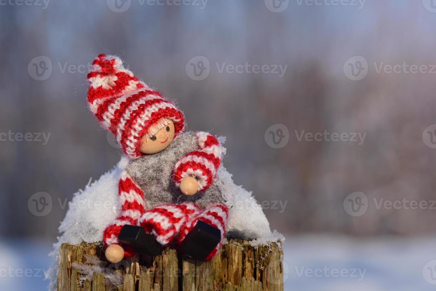 Close up of little doll with winter clothes and hat sitting on a wooden post in the snow photo