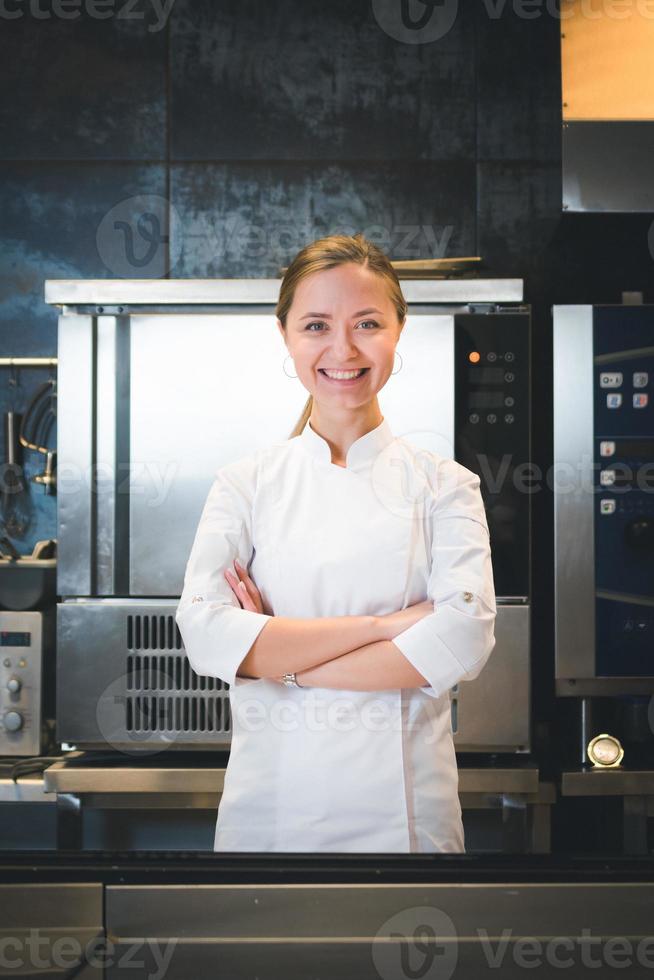 Portrait of confident and smiling young woman chef dressed in white uniform, professional kitchen are on background photo
