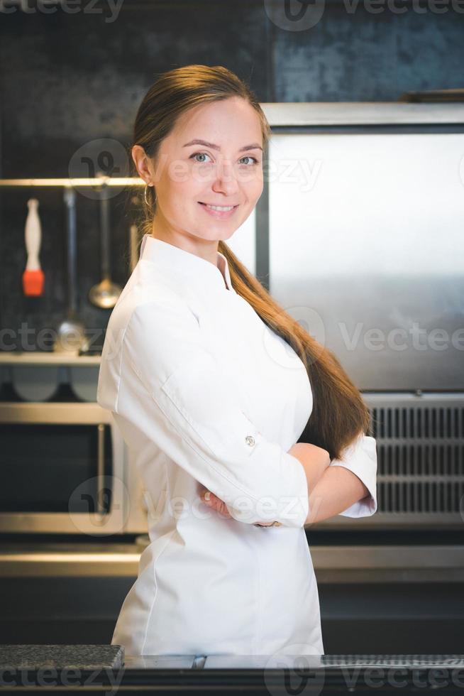 retrato de una joven chef confiada y sonriente vestida con uniforme blanco, la cocina profesional está en segundo plano foto