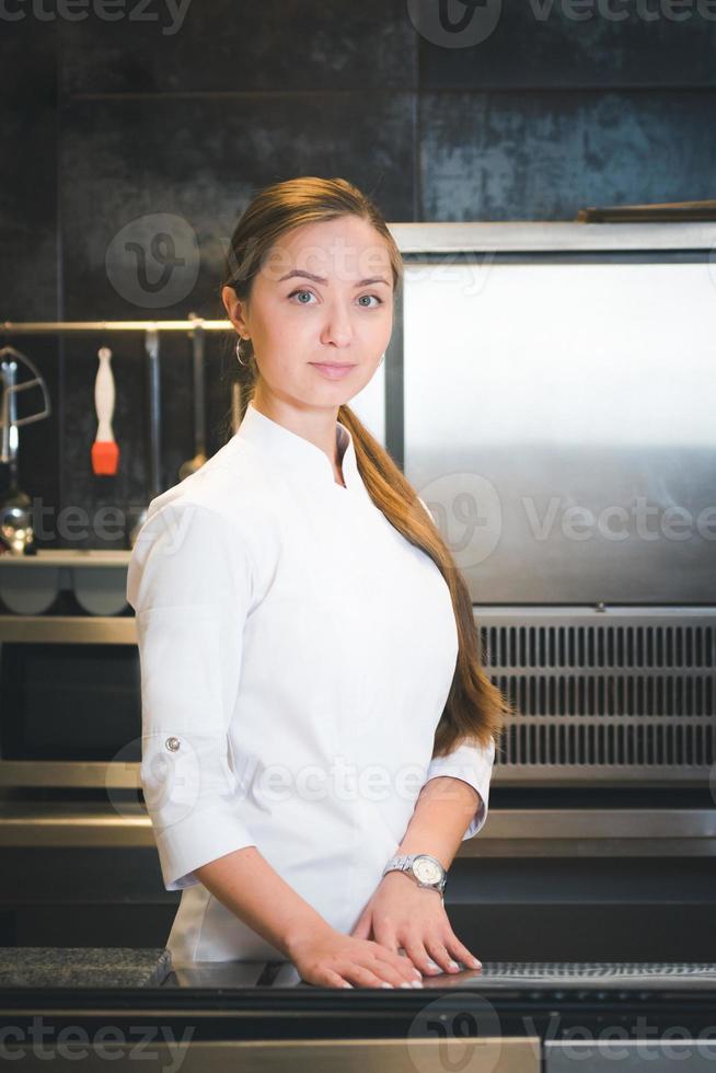 Portrait of confident and smiling young woman chef dressed in white uniform, professional kitchen are on background photo