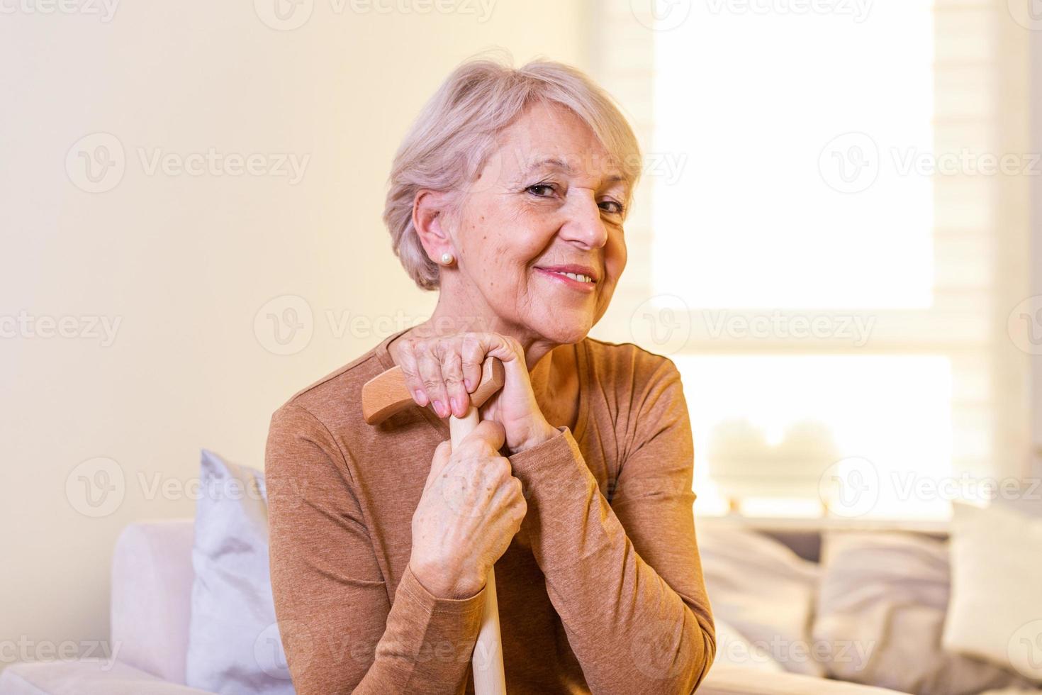 retrato de una hermosa anciana con cabello blanco y bastón. retrato de una anciana sentada en un sofá en casa. Sonriente mujer madura de pelo gris de mediana edad mirando a la cámara foto