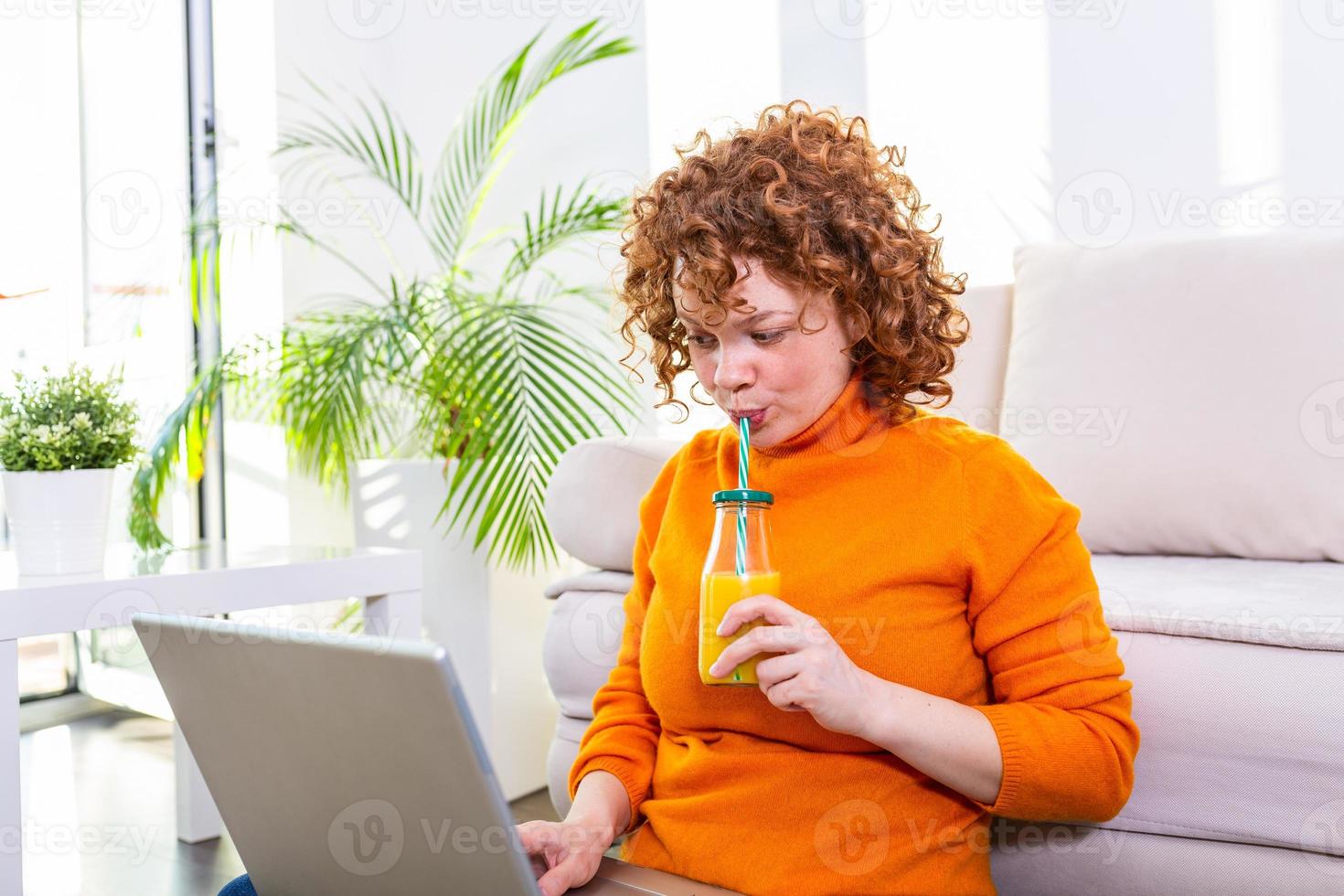 Cute female red hair freelancer reading notebook while sitting on floor with open computer in modern home, young creative woman work on laptop while having orange juice photo