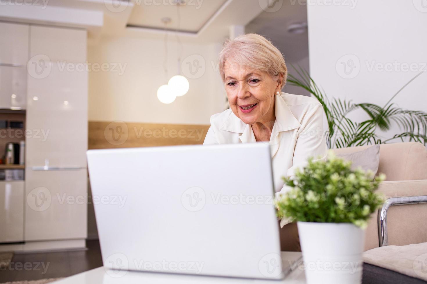 Portrait of senior woman sitting at table at home and working on her laptop. Older lady surfing the net from home while sitting on her sofa and using laptop computer photo