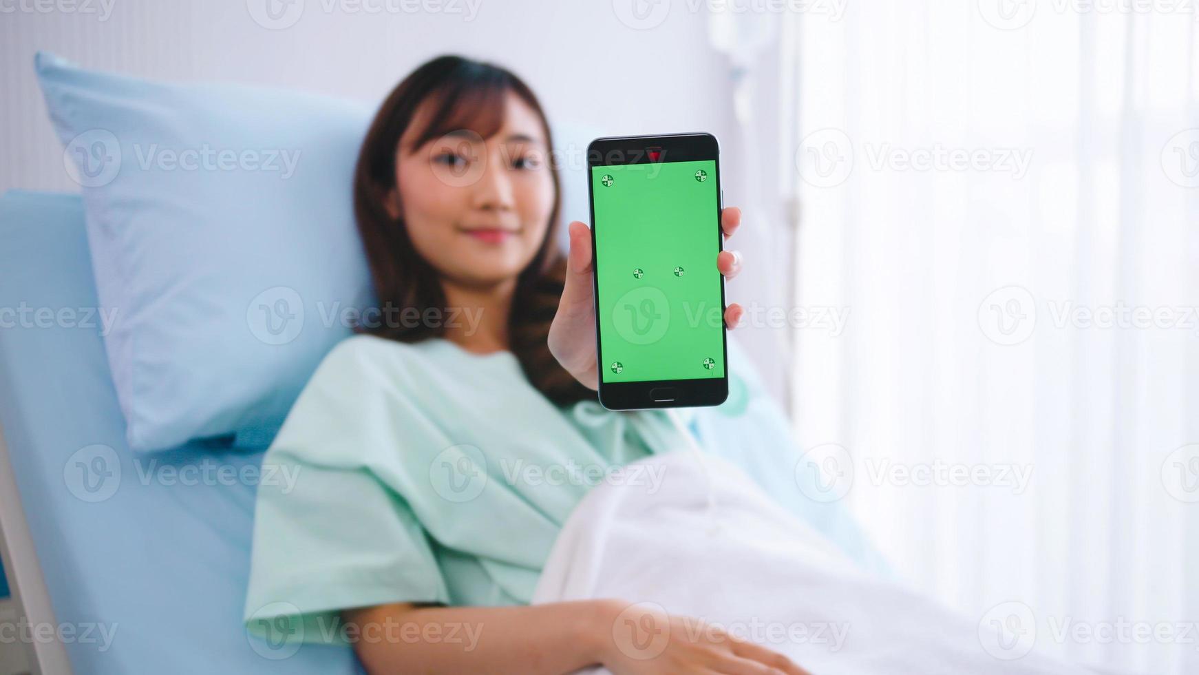 feliz joven paciente asiática que muestra un teléfono inteligente con pantalla verde durante la hospitalización. foto