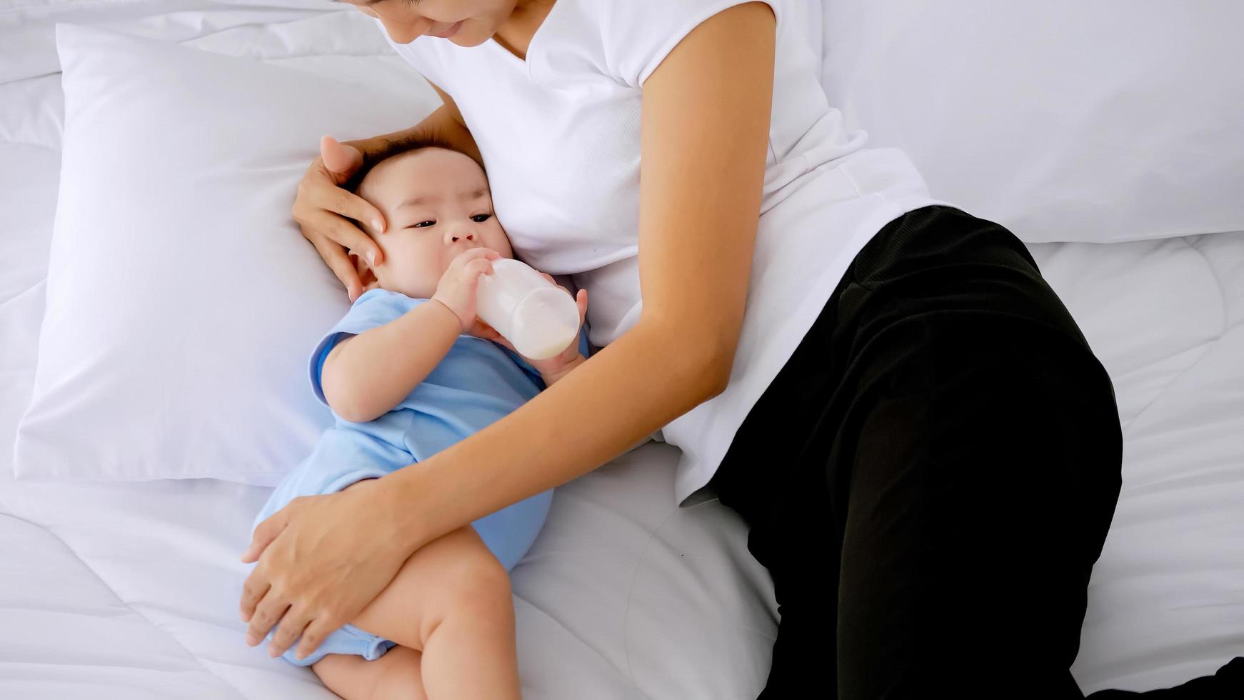 joven madre alimentando al pequeño bebé de la botella de leche en la cama. foto