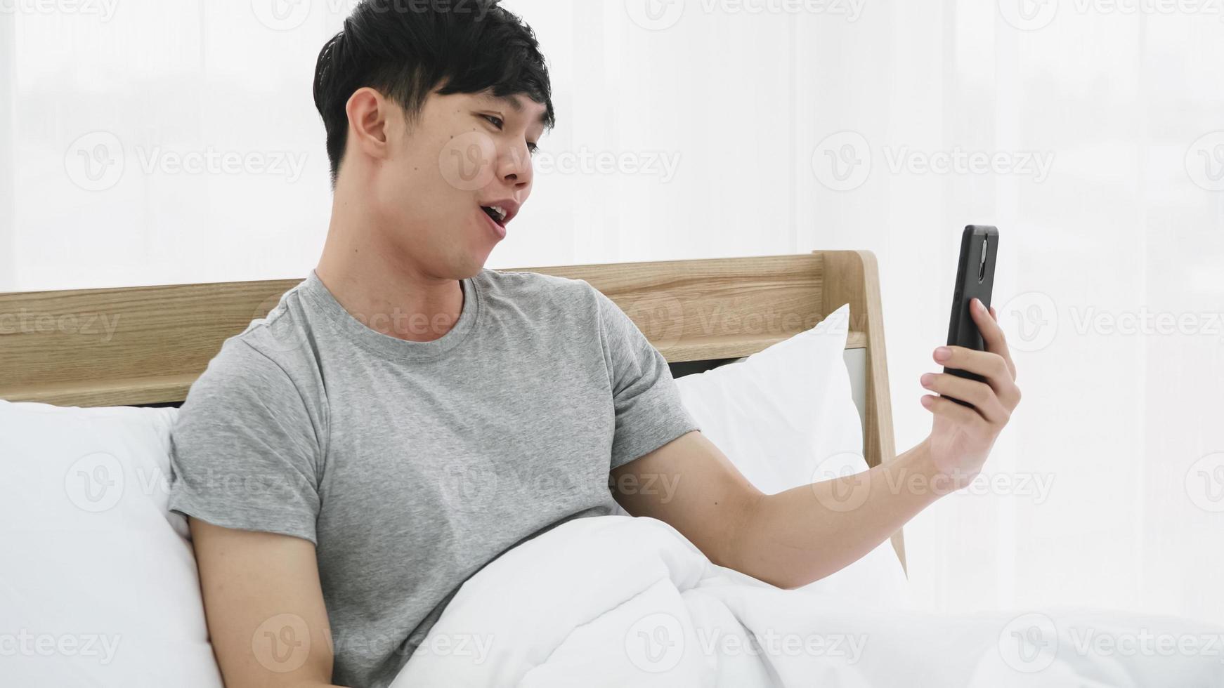 Young happy man using mobile phone for video call in bed at home photo