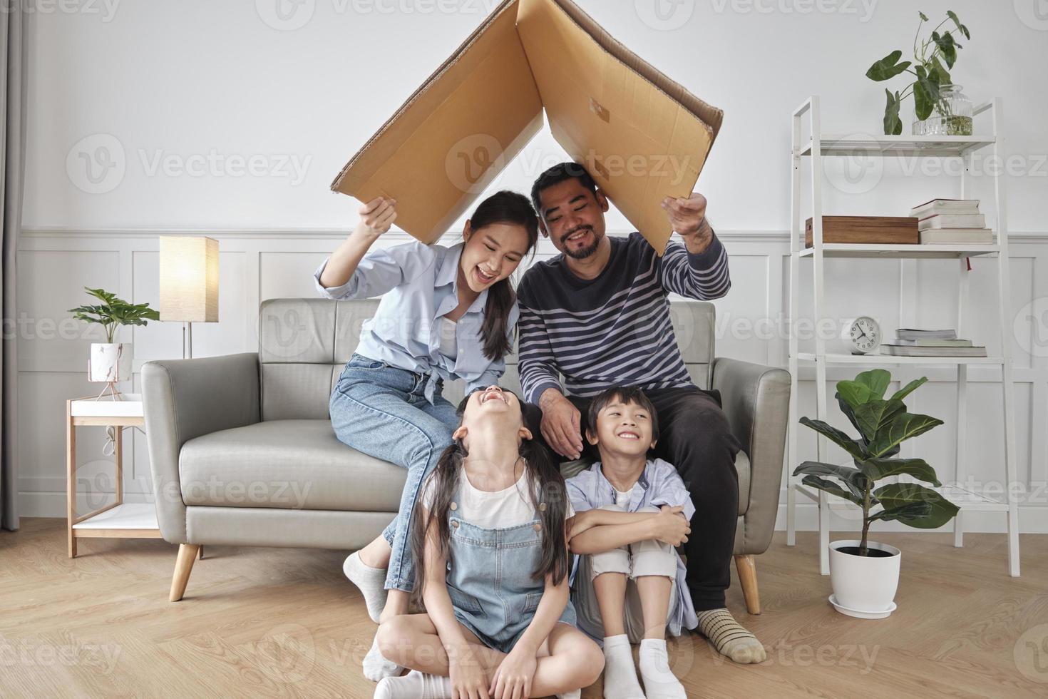 Asian Thai family, adult dad, mum, and little children happily fun playing in a white living room, built cardboard paper safety house with imagination, lovely weekend and wellbeing domestic lifestyle. photo