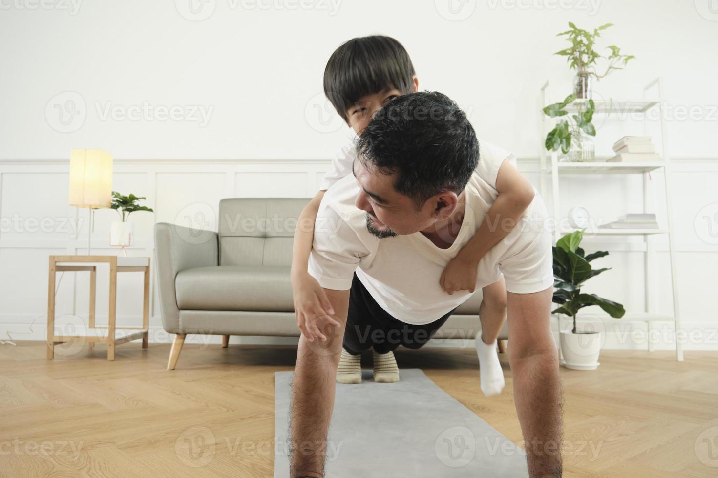 Asian Thai father and son are fitness training exercise and practices yoga on living room floor, lovely teasing together for health and wellness, happy domestic home lifestyle on family weekend. photo