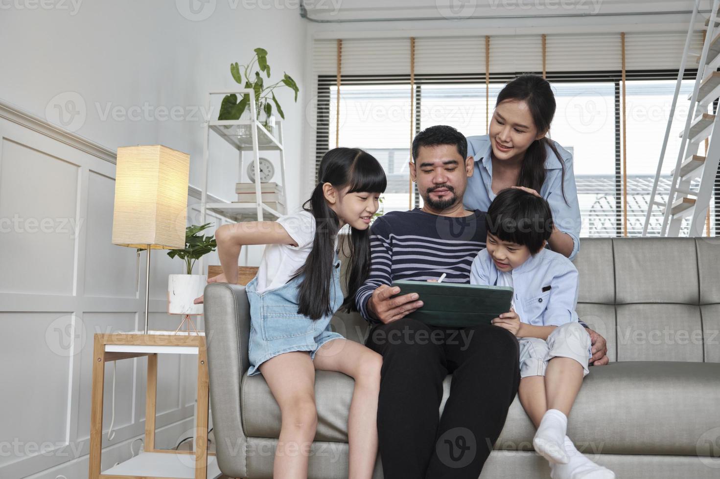Happy Asian Thai family, parents, and children have fun using digital tablet together on sofa in home living room, a lovely leisure weekend, and domestic wellbeing lifestyle with internet technology. photo