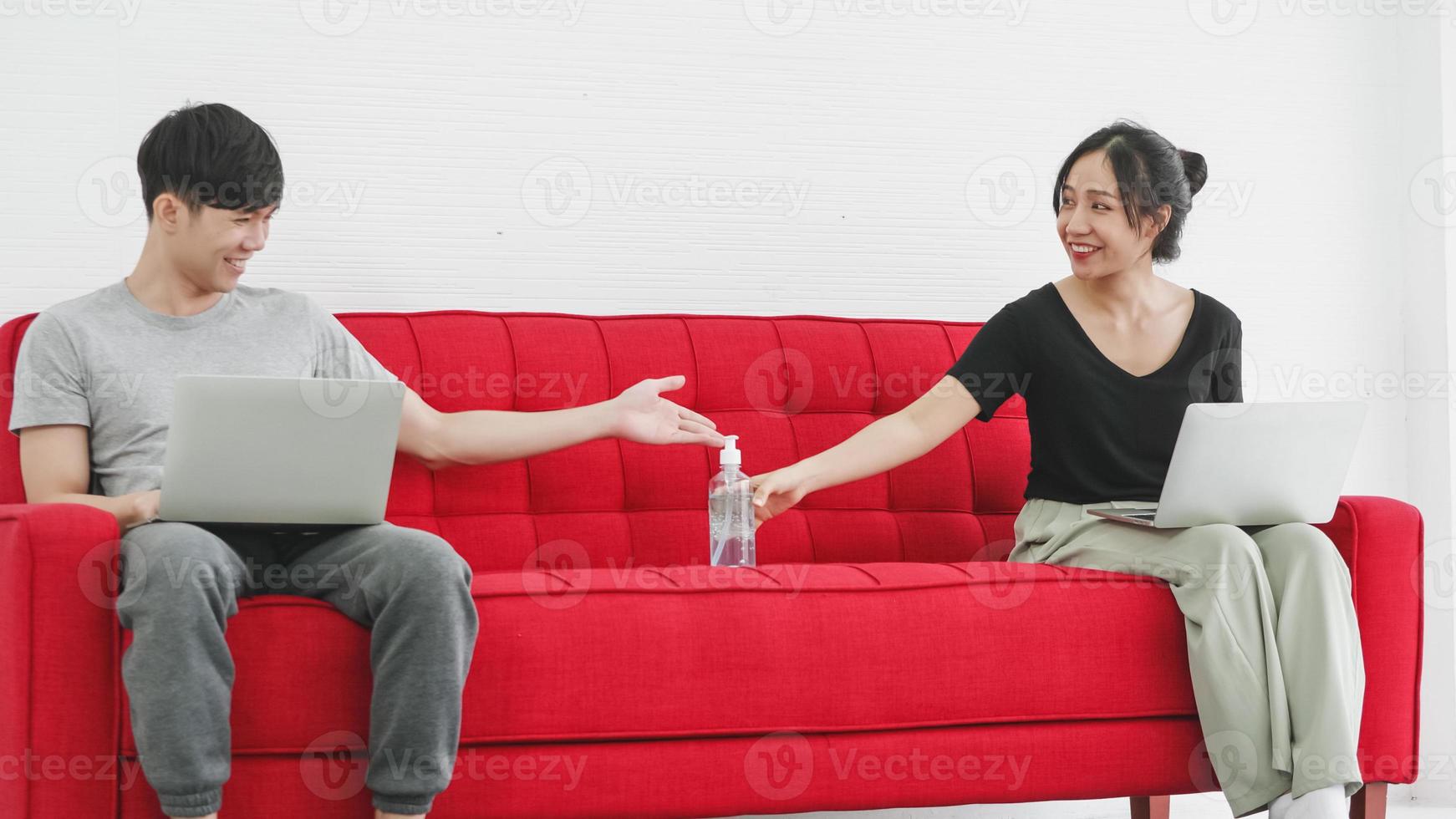 Happy Asian couple cleaning hands with sanitizer alcohol gel while sitting working together on sofa at home. photo
