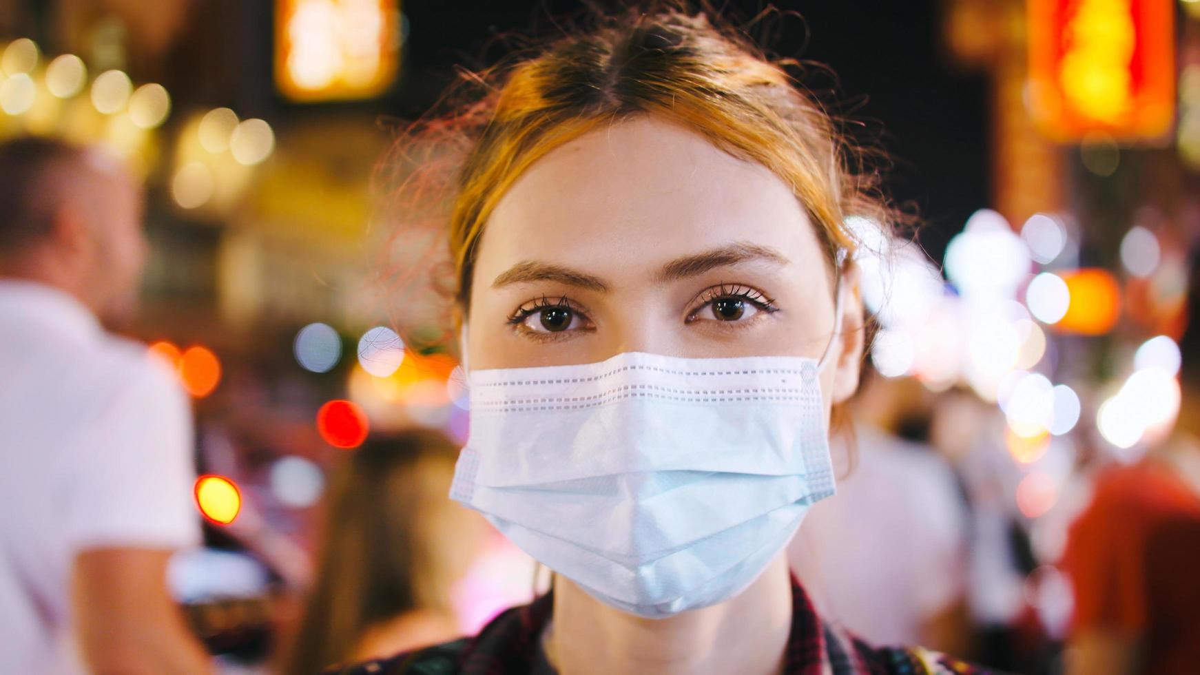 Portrait of a tourist woman wearing medical mask while standing on the street of city at night. photo