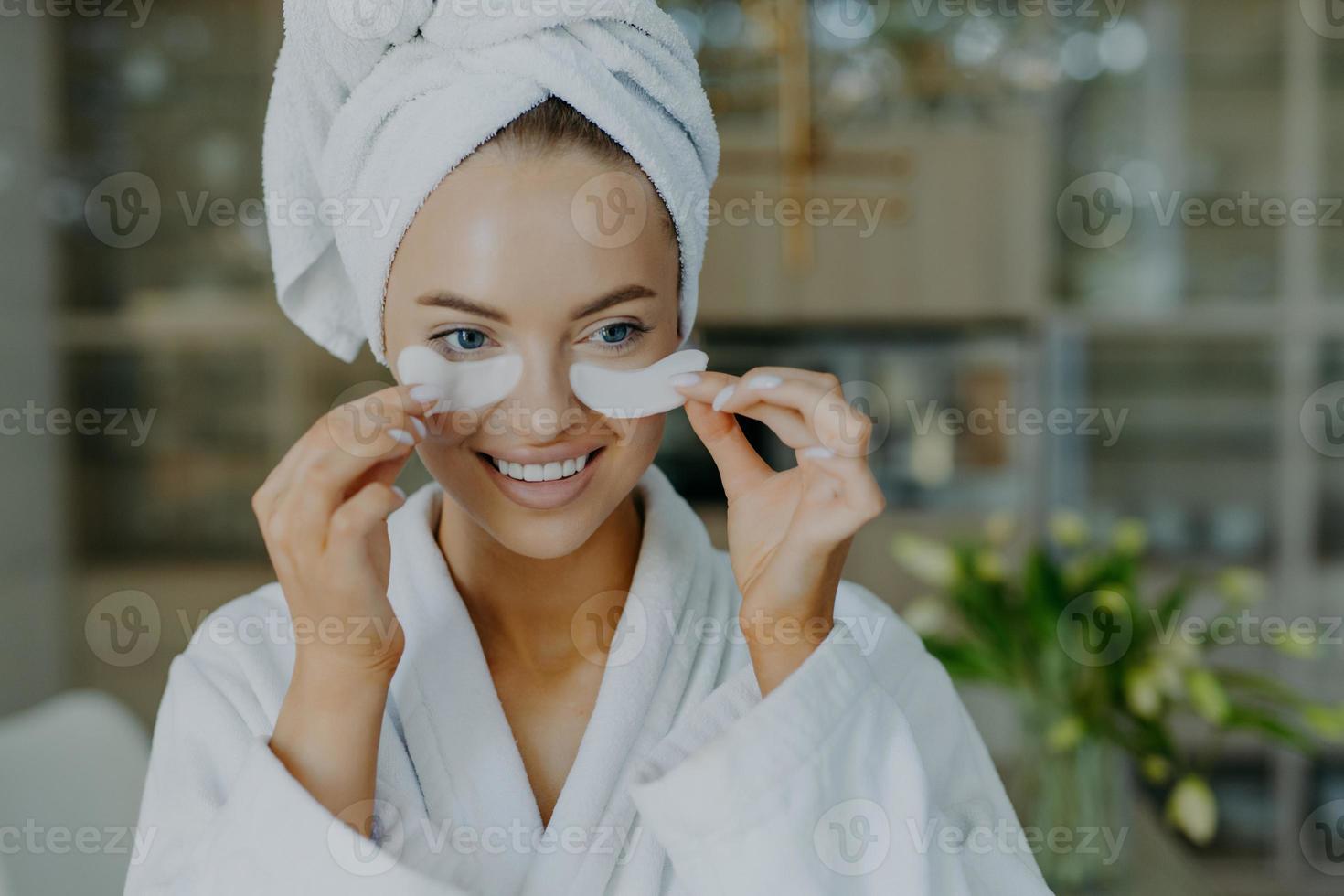 Close up shot of happy lovely European woman takes off beauty patches smiles gently has refreshed well groomed complexion wears bathrobe and wrapped towel undergoes cosmetic procedures at home photo