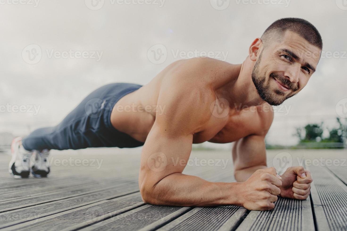 Workout and healthy lifestyle concept. Pleased muscular bearded young man stands in plank pose, makes sport training outdoors, looks with determined expression, concentrated on getting better photo