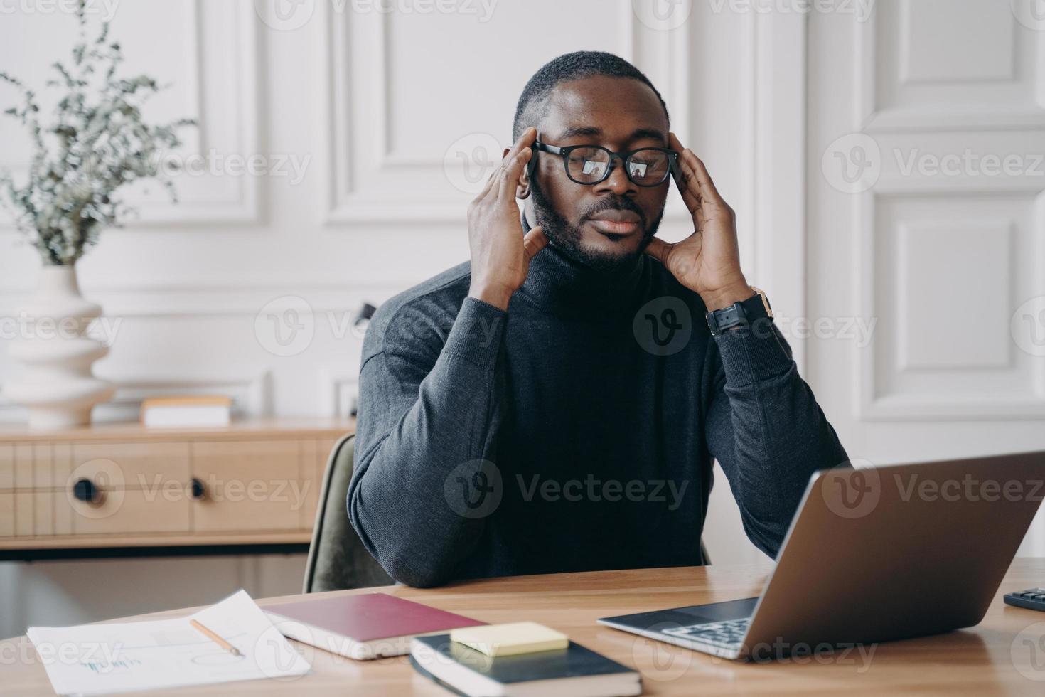 African male bookkeeper sits at desk in front of laptop eyes closed fingers gently touching temples photo