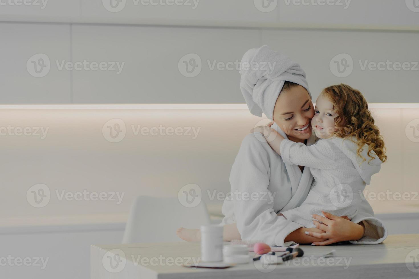 Young mom and little daughter in white bathrobes hugging while doing hygiene or spa procedures together photo