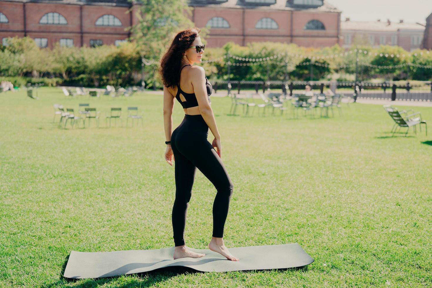 Una mujer morena en forma vestida con ropa deportiva se para descalza en karemat tiene un entrenamiento fino en el césped verde al aire libre durante el verano usa gafas de sol parece estar en buena forma física en algún lugar foto