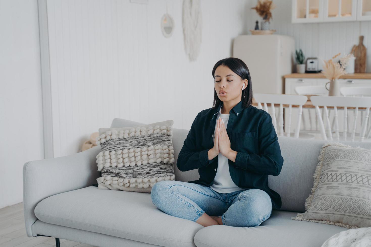 Calm woman breathing practicing yoga sitting in lotus pose on couch at home. Wellness, stress relief photo