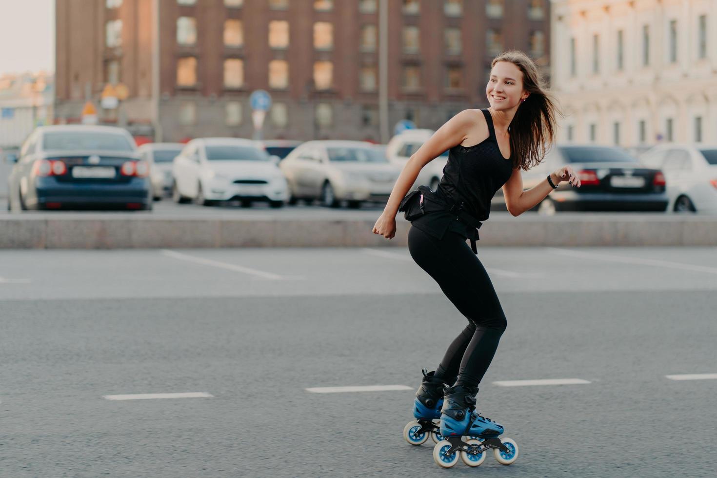 Outdoor shot of slim cheerful woman dressed in active wear looks away leads healthy lifestyle rides on rollers poses on road in urban place feels energized. Rollerblading as hobby. Recreation photo