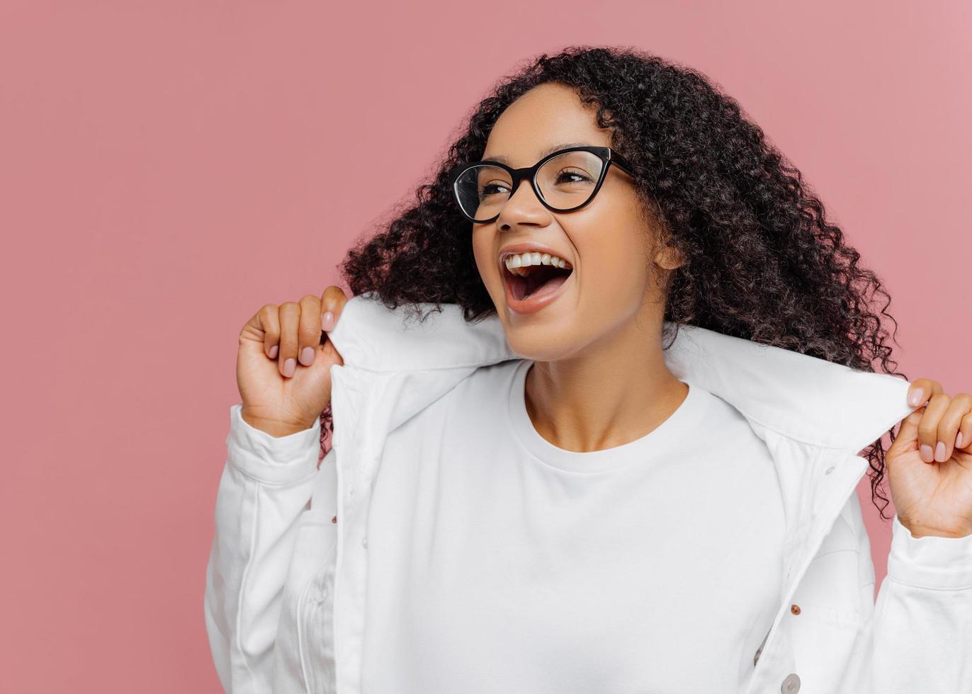 Curly overemotive woman laughs happily, keeps hands on collar of white jacket, has healthy skin, wears spectacles, curly hair, looks on left side isolated on pink background. People, fashion, emotions photo