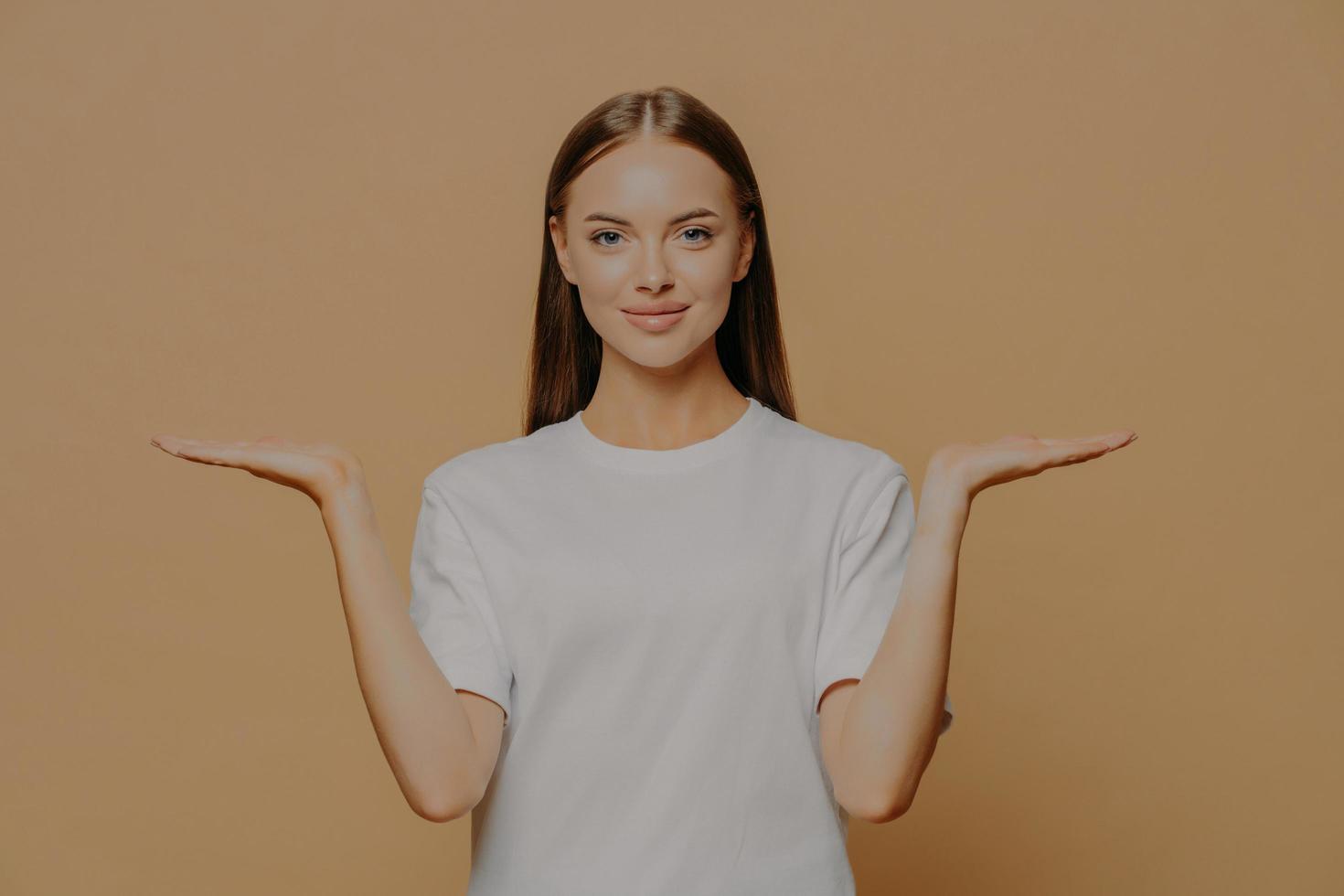 Beautiful dark haired European woman raises palms pretends holding something over blank copy space against brown background wears casual white t shirt proposes product. Advertisement gesture photo