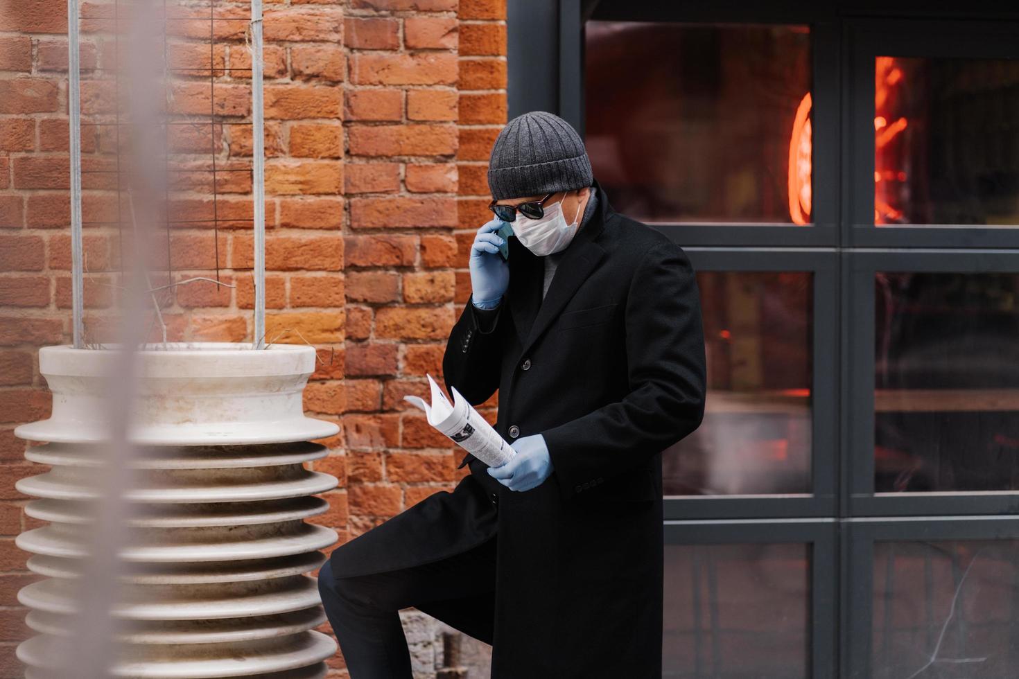 Male entrepreneur discusses working issuses via smartphone, wears protective medical mask and gloves, holds papers, poses brick wall. Epidemic virus. Novel coronavirus concept. Stay safe and healthy photo