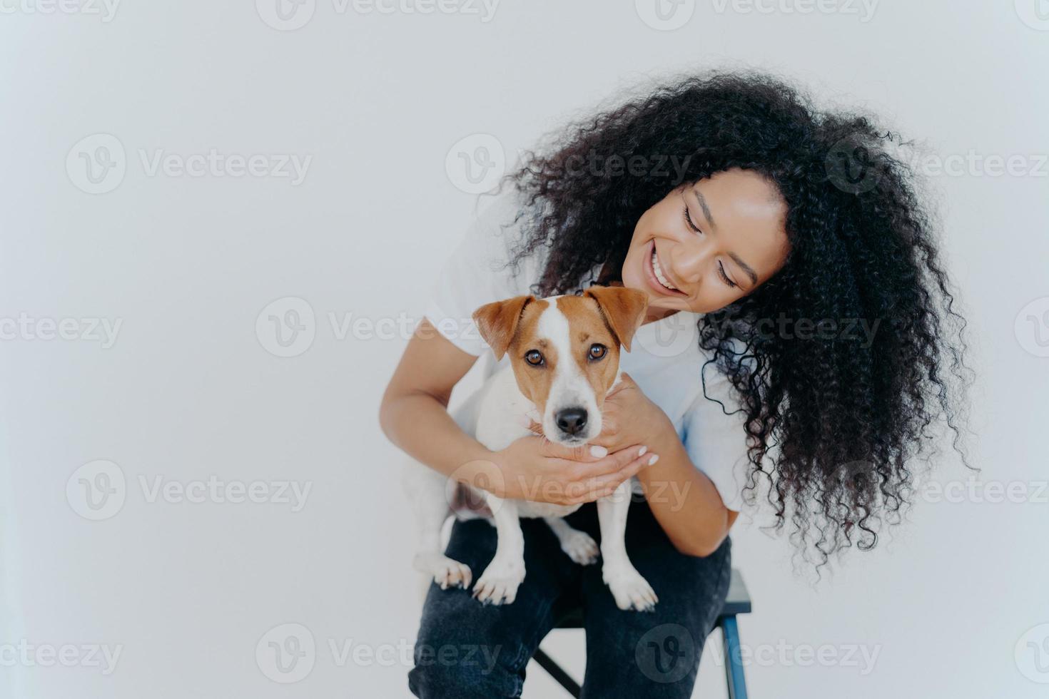 Glad dark skinned girl plays with jack russell terrier dog, have fun together, poses against white background, dressed casually. Happy Afro girl poses with pet indoor. People, animals, fun concept photo