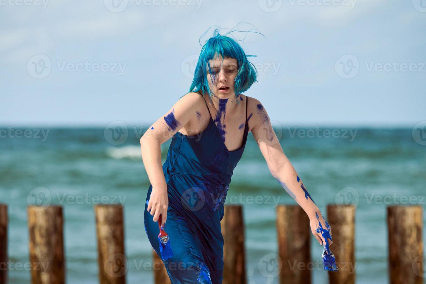 Artistic blue-haired woman performance artist smeared with blue gouache paints dancing on beach photo