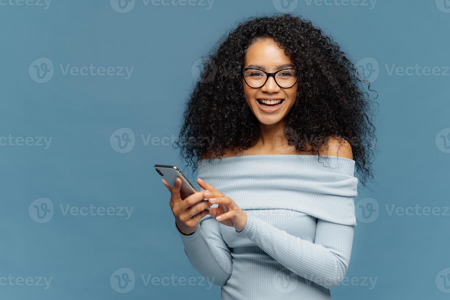 Lovely teenage girl enjoys online communication, holds modern cell phone, checks email box, connected to wireless internet, cannot imagine life without technologies, isolated on blue studio wall photo