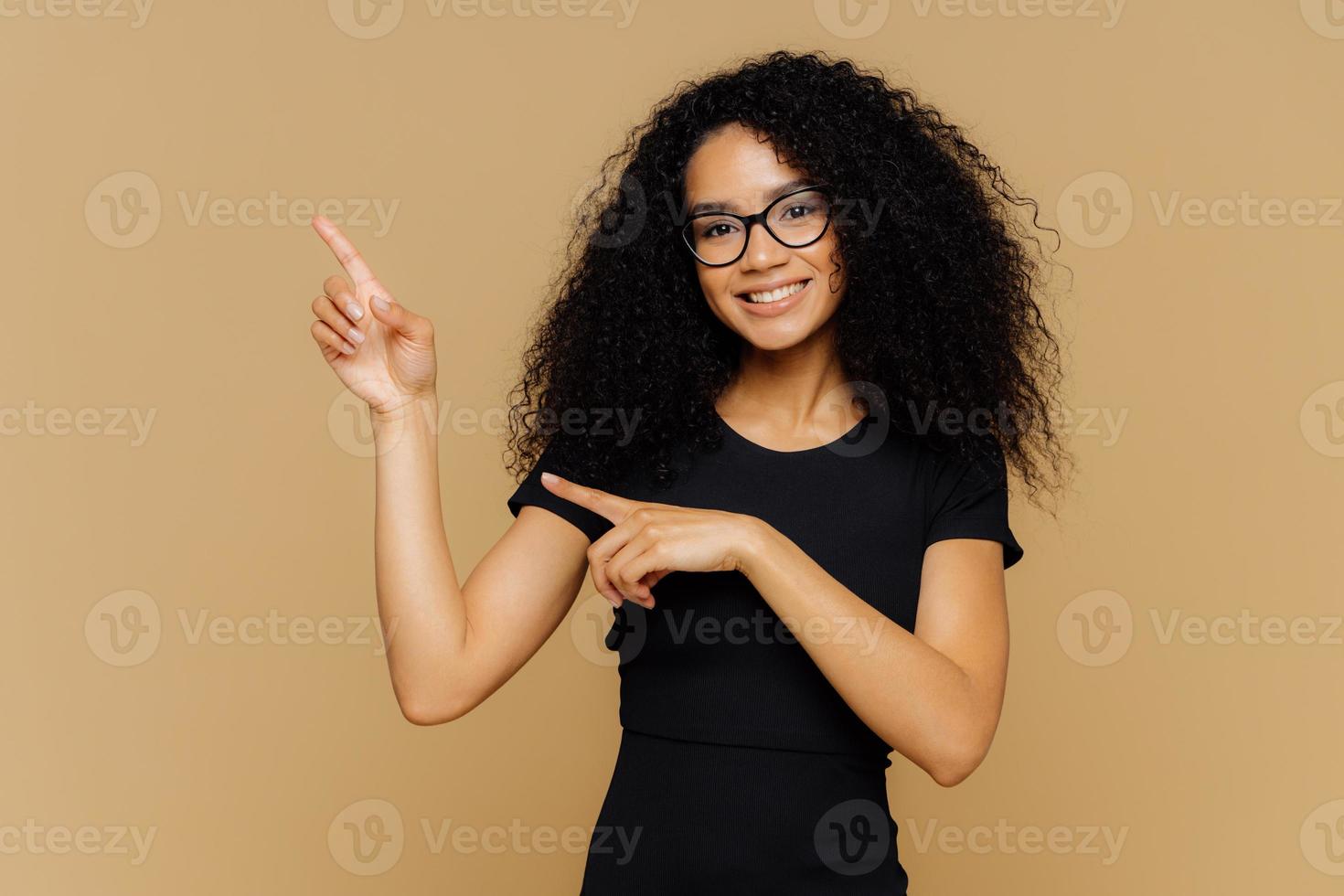 una foto aislada de una mujer afroamericana feliz apunta hacia arriba y hacia un lado, demuestra un espacio en blanco para su anuncio, usa anteojos, una camiseta informal, tiene una sonrisa amable en la cara. gente y promoción