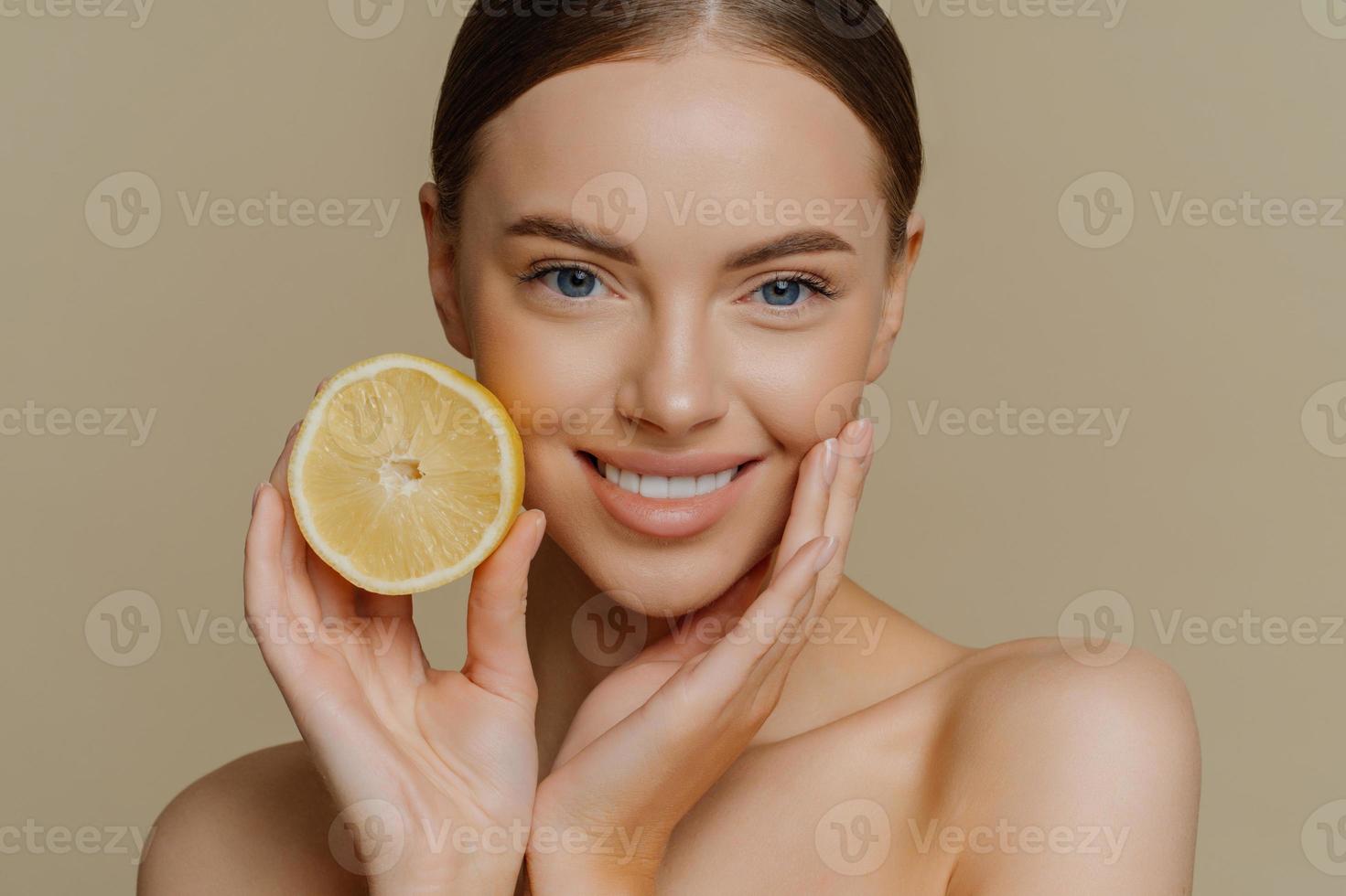 Indoor shot of beautiful dark haired gentle woman smiles pleasantly poses with half of lemon for skin and body care poses bare shoulders indoor isolated over brown background. Cosmetology concept photo