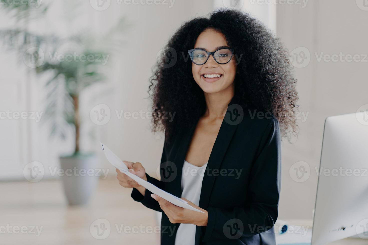 Photo of happy female entrepreneur with Afro hairstyle, studies documentation, wears spectacles and elegant clothes, stands in office interior, prepares to present her business ideas for colleagues