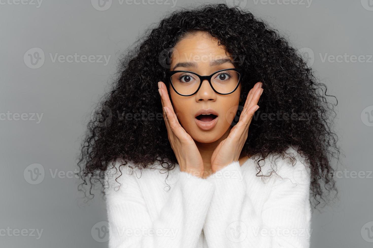 Omg, it cannot be so Surprised emotional dark skinned young female with curly hair, touches both cheeks, has shocked faciall expression, dressed in white jumper, isolated over grey background photo