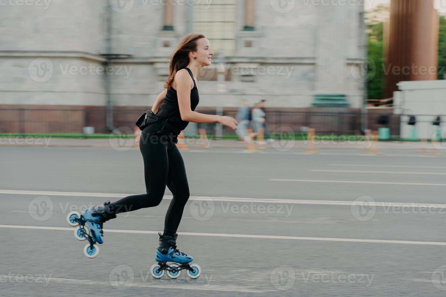 Summer rollerblading and fitness concept. Fit young dark haired woman fond of rollerskating exercises all body poses on road along asphalt during sunny day wears comfortable clothes has sporty figure photo
