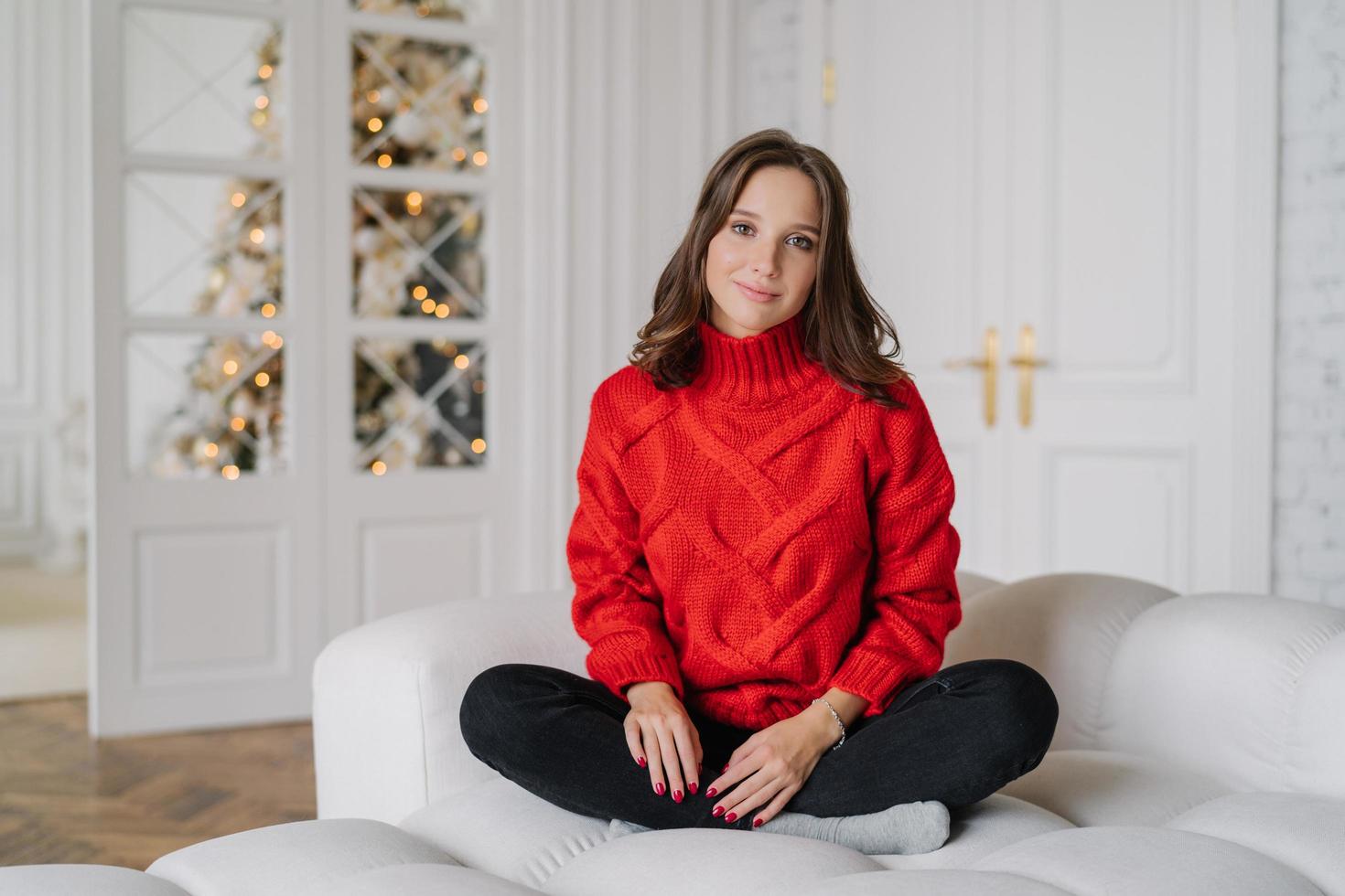 Photo of calm carefree girl wears warm doemstic clothes, poses on white couch, looks directly at camera, spends winter weekend at home, anticipates New Year, decorated fir tree. Holidays concept