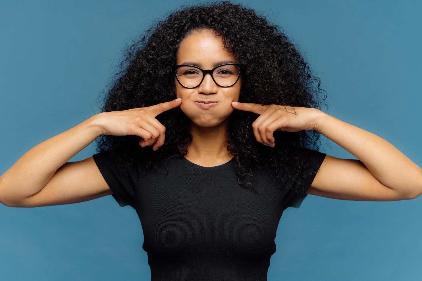 Photo of funny merry female blows cheeks, touches with both index fingers, wears black casual t shirt, optical eyewear, stands over blue background, makes grimace and foolishes indoor alone.