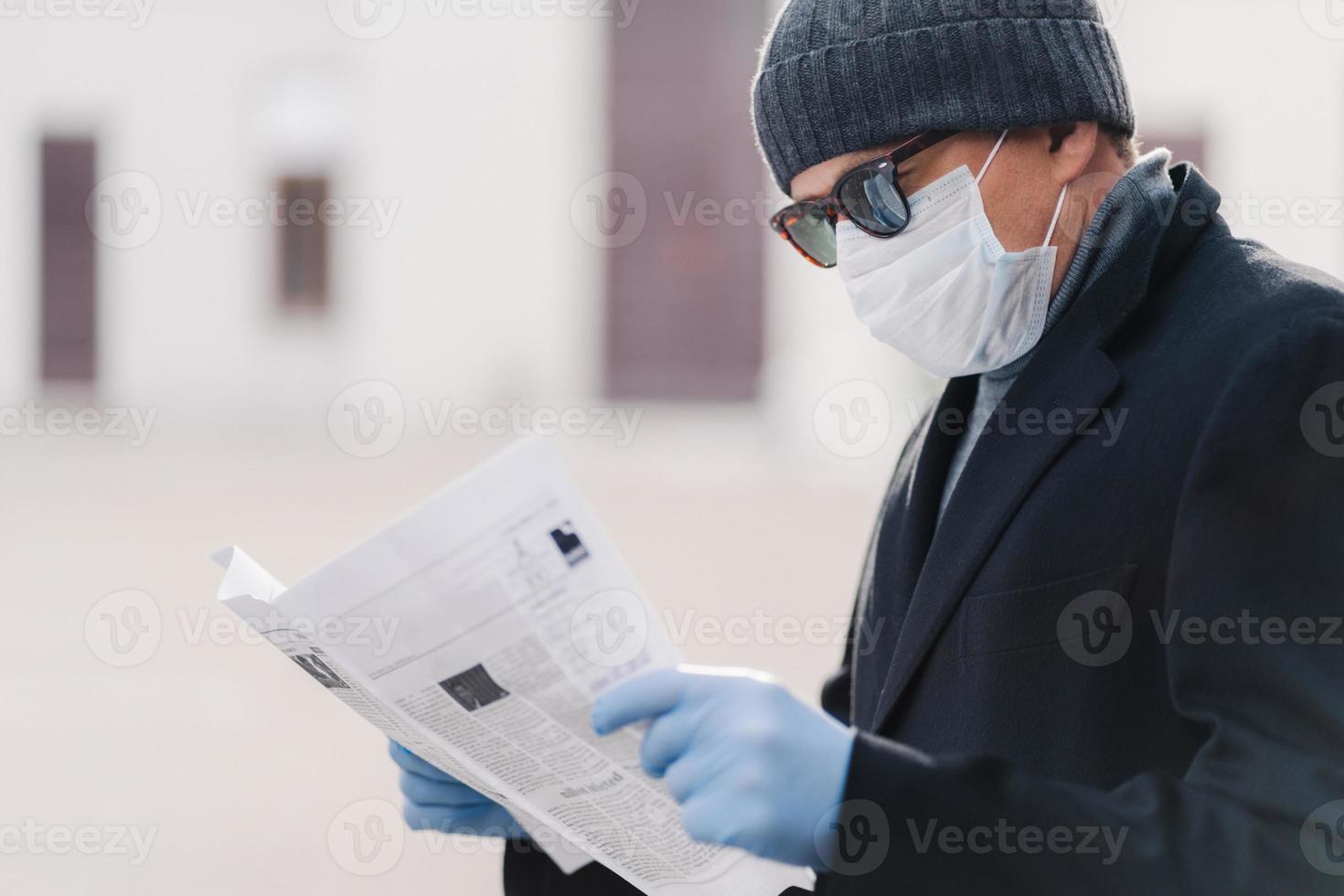 Quarantine for coronavirus. Horizontal shot of adult man wears protective mask and disposable gloves, reads fresh newspaper, finds out news about pandemic situation. Infection prevention concept photo