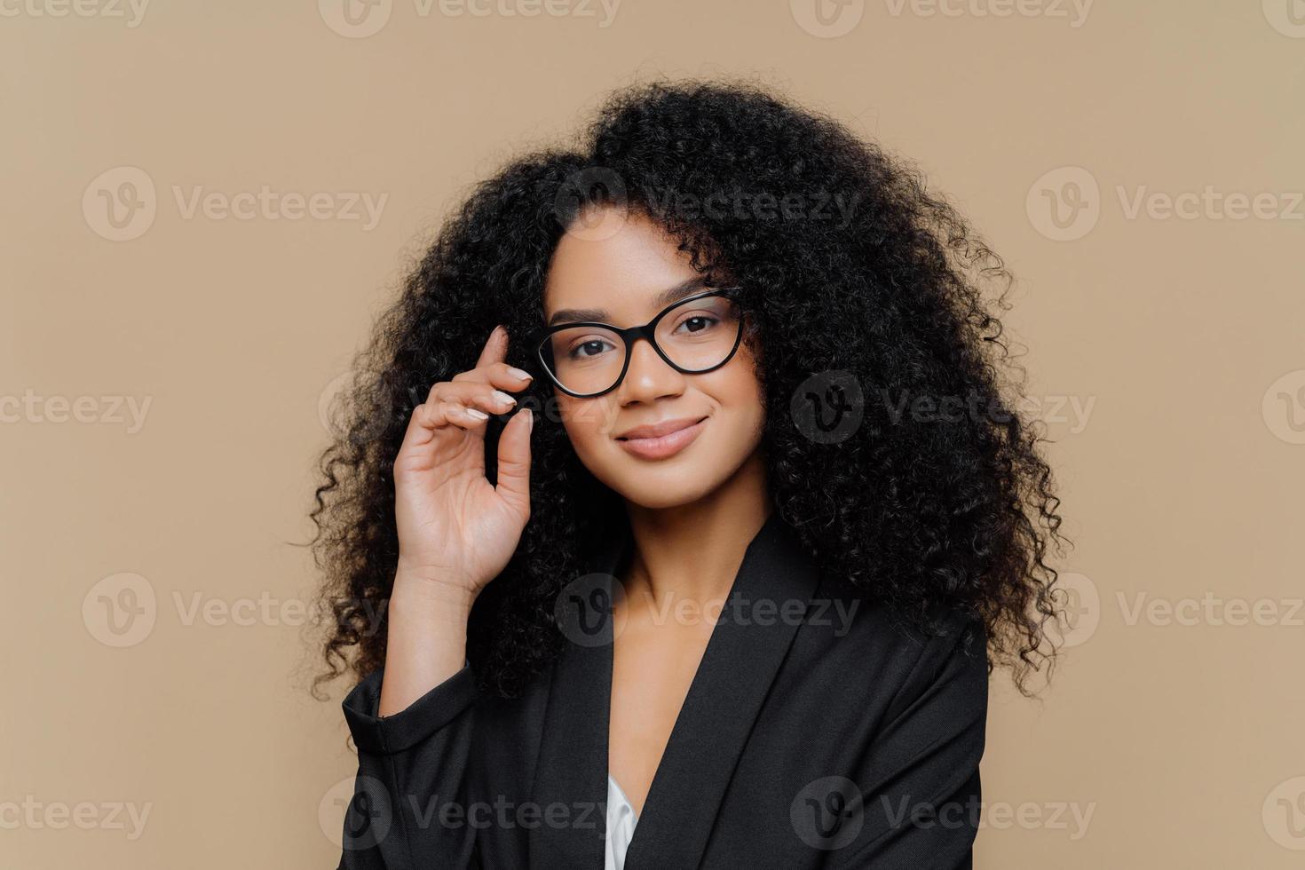 Close up portrait of calm satisfied Afro American woman with bushy curly hairstyle, keeps hand raised, has healthy skin, wears spectacles, black elegant outfit isolated on brown wall. Face expressions photo