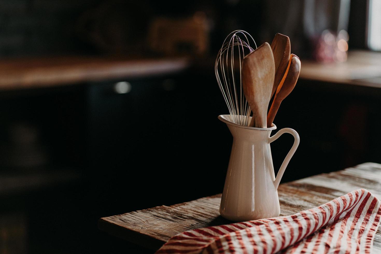 Kitchen accessories on wooden table. Utensils in white ceramic jar against dark background. Rustic style. Dishware for preparing meal. Wooden spoons shpatula and whisk photo