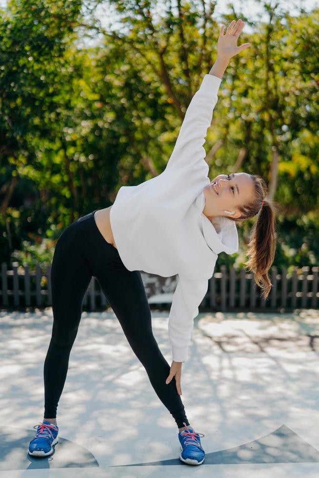 Slim healthy woman exercises outdoor keeps arm raised stretches body before running enjoys physical activity dressed in active wear breathes fresh air listens music from playlist. Sport concept photo