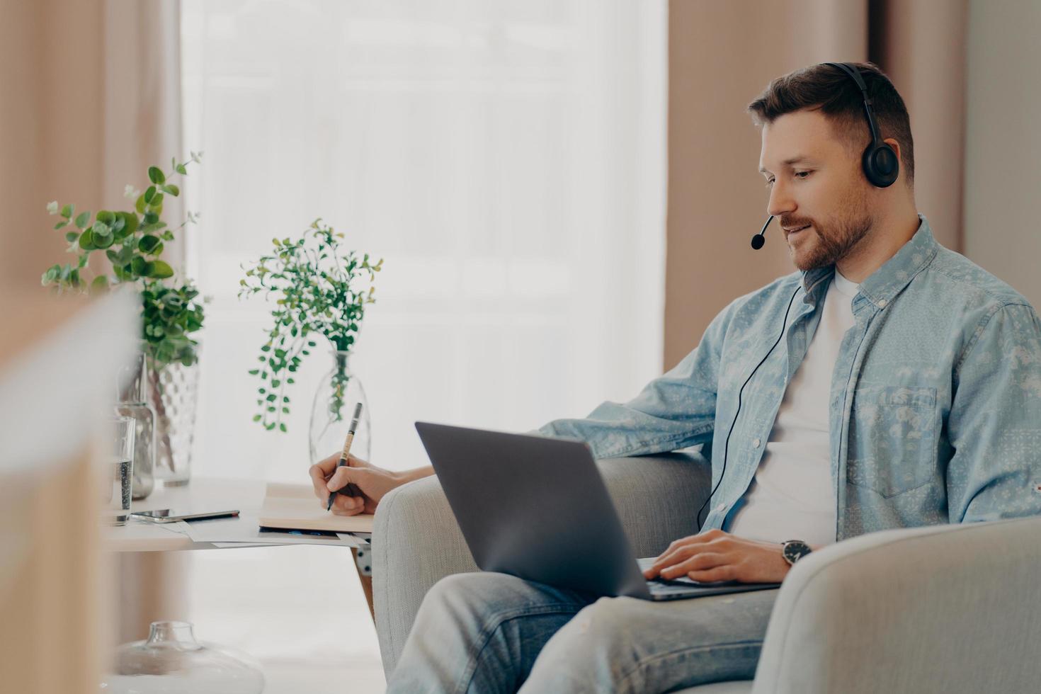 Male freelancer listens attentively tutorials how to start own business writes down information wears stereo headphones and laptop computer works online from home makes video call watches webinar photo