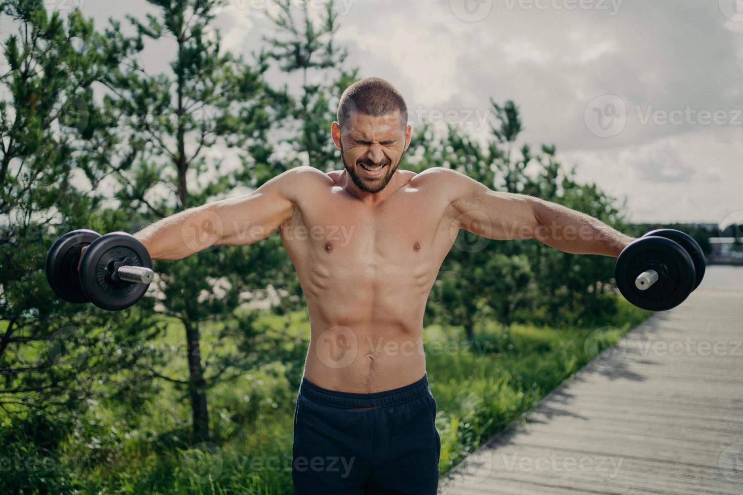 Powerful muscular sportsman demonstrates endurance, lifts heavy barbells and trains arms muscles, poses shirtless outdoor, puts all efforts in lifting weight. Athletic man works out with dumbbells photo