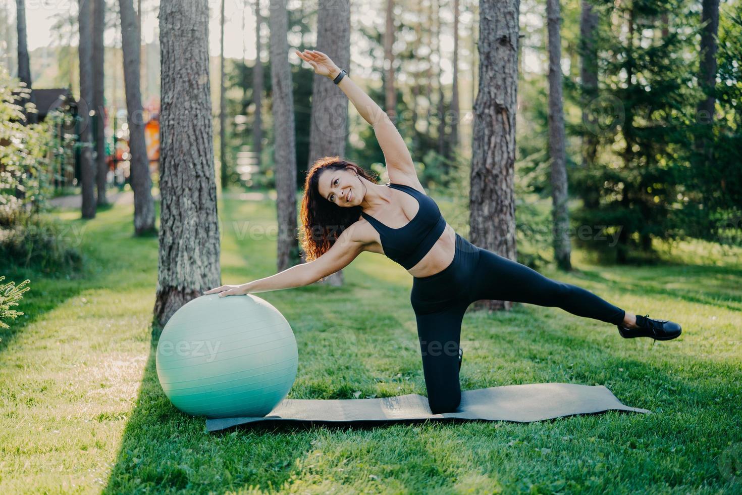 Horizontal shot of active slim woman with cheerful expression leans aside does exercises with fitness ball, dressed in sportsclothes, poses outdoor against tall trees on karemat. Healthy lifestyle photo