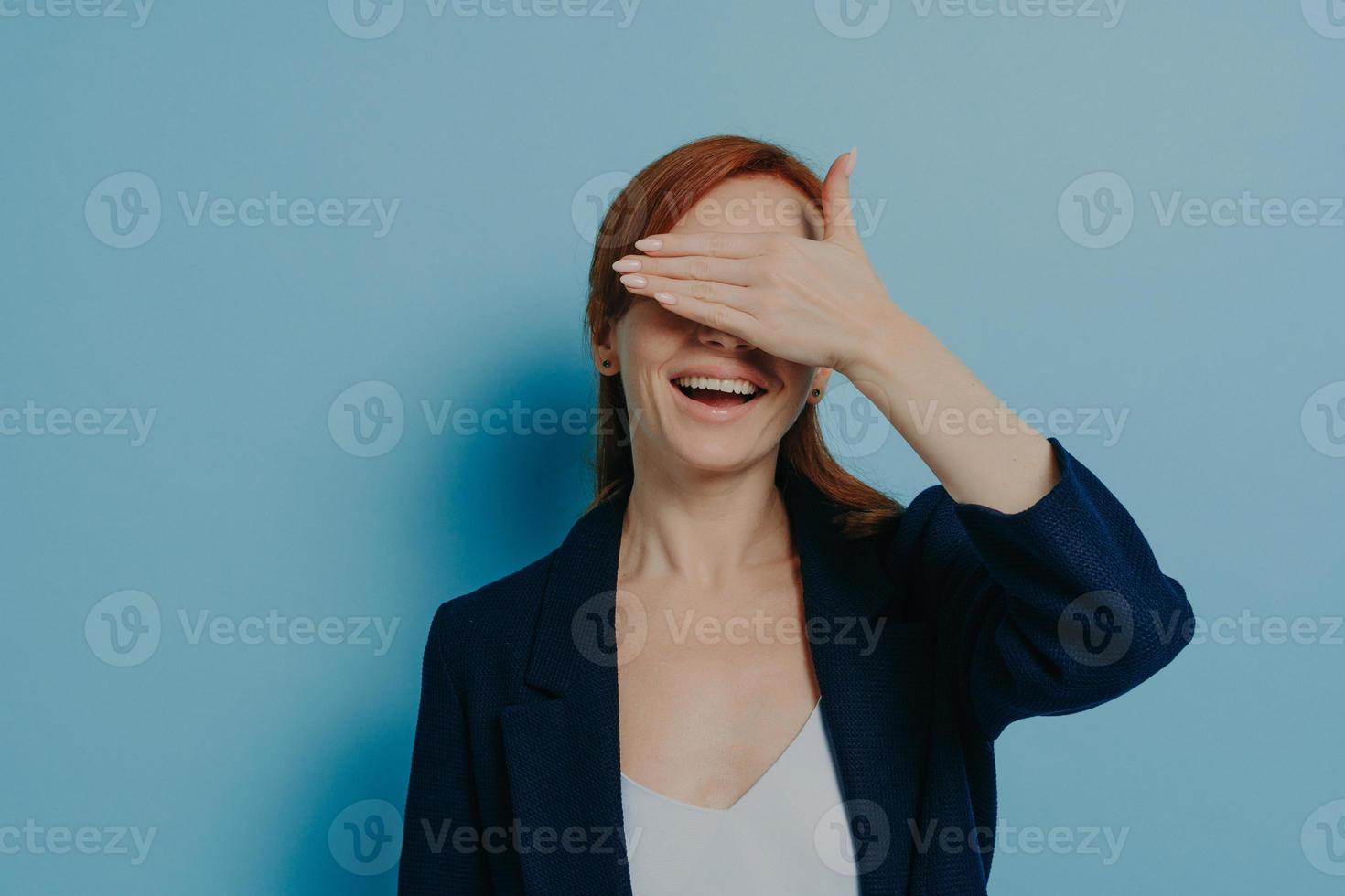 juguetona y graciosa chica pelirroja cubriendo sus ojos con palma aislada en fondo azul, aislada en azul foto