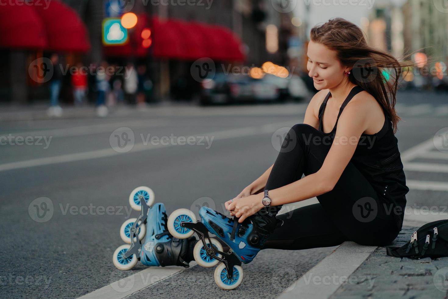 Outdoor activities fit lifestyle. Sideways shot of active woman sits on road adjustes rollerblades prepares for skating puts on inline skates has hair floating on wind enjoys weekend favorite hobby photo