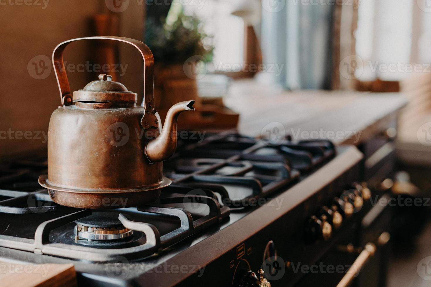 Old aluminium kettle boilng on gas stove in kitchen against cozy blurred background. Antique item made of copper metal. Vintage style photo