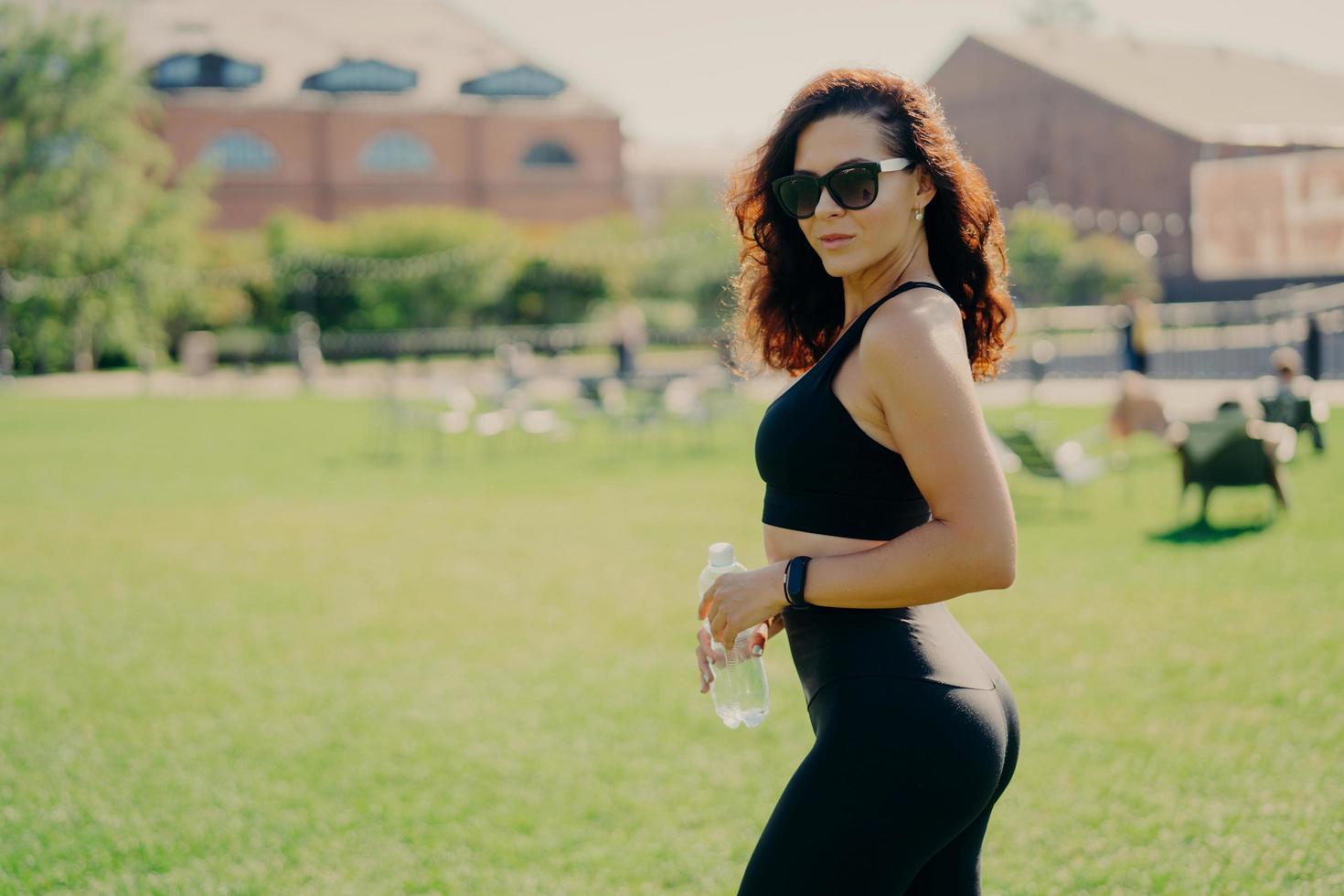 Summer activity and energy concept. Brunette sporty woman in sports clothes holds bottle of water wears sunglasses poses outdoor poses against blurred background with green lawn. Sporty lifestyle photo