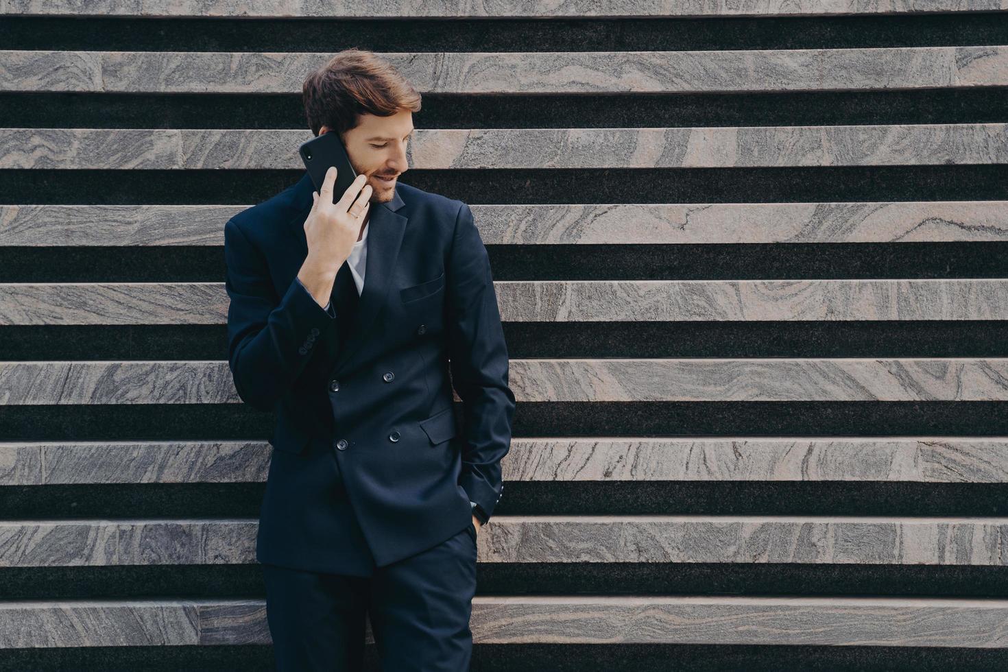 Businessman in elegant black clothes phones to partner focused down keeps smartphone photo