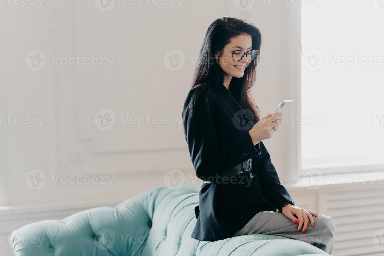 European entrepreneur wears corporate suit and spectacles, uses online banking application for transferring money, holds cellular, sends email, poses at back of comfortable sofa, uses free wifi photo
